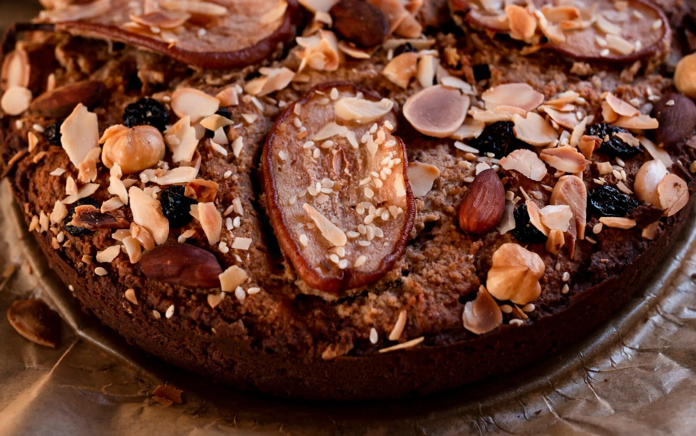 Close-up of a rustic date cake topped with sliced almonds, hazelnuts, sesame seeds, and dried fruits on a baking sheet.