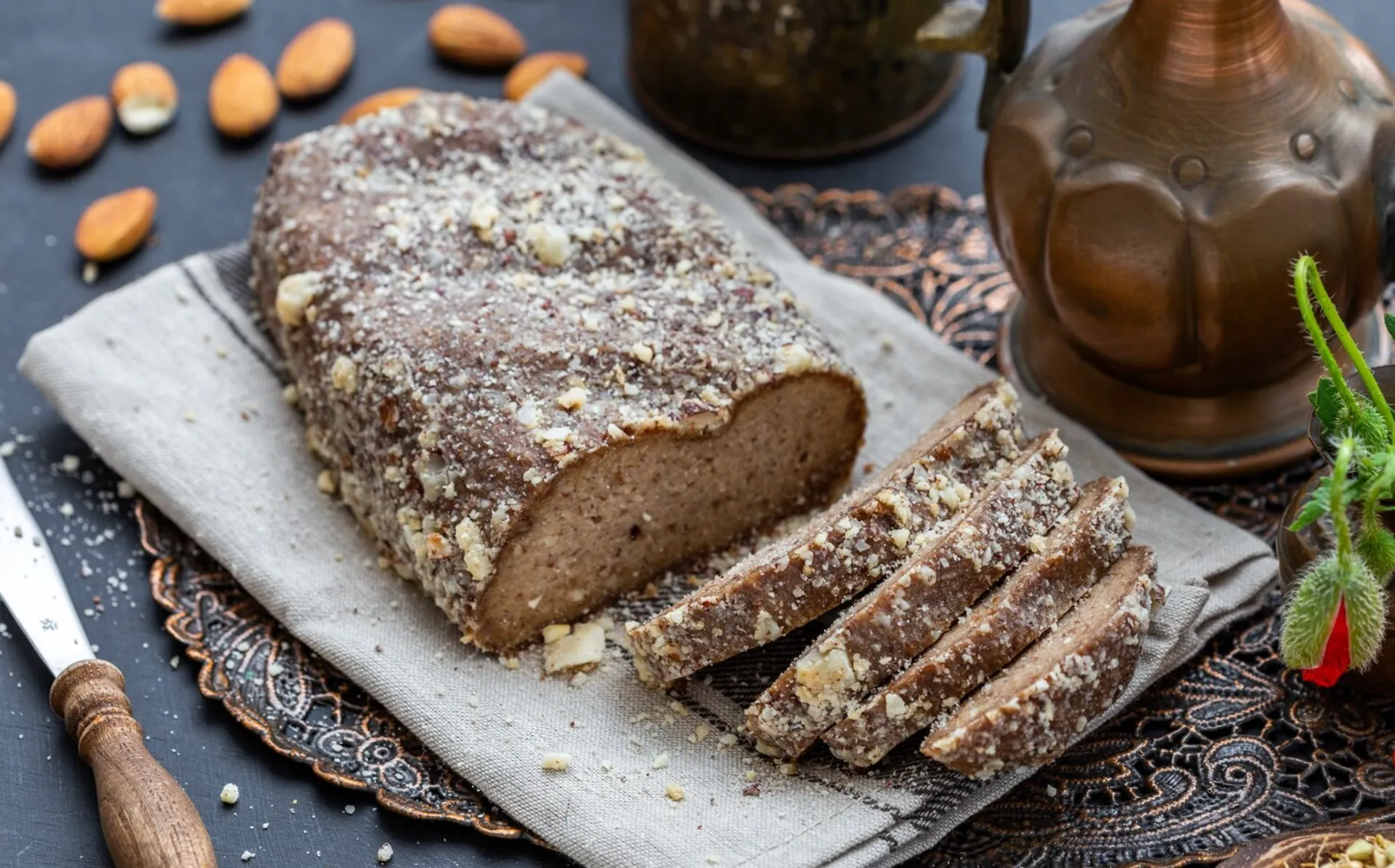 A freshly baked loaf of date nut bread, sprinkled with chopped nuts, placed on a linen cloth with decorative serving ware in the background.