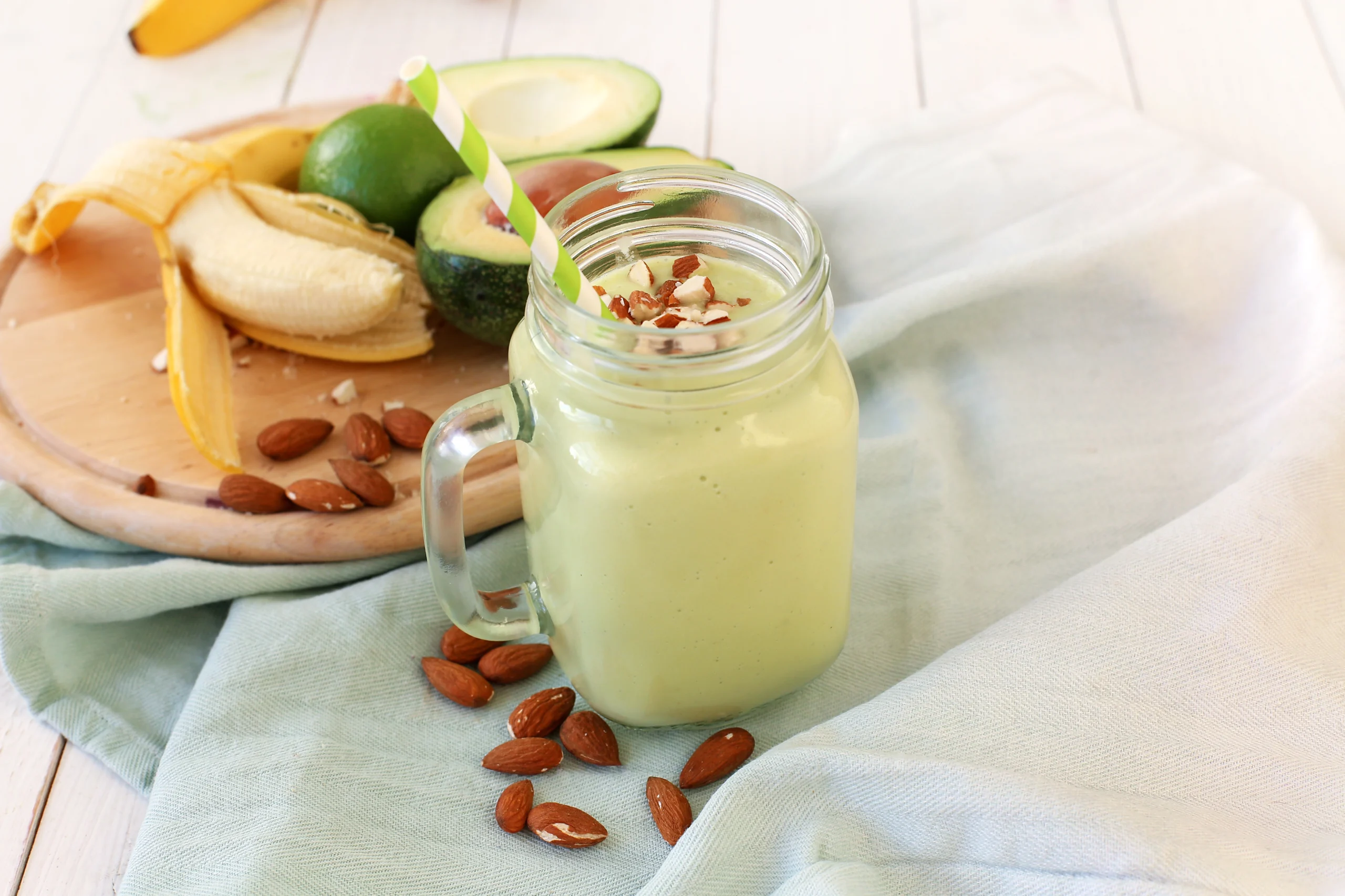 A creamy avocado and banana smoothie served in a glass jar with a green striped straw, topped with chopped almonds. Fresh avocados, bananas, lime, and whole almonds are displayed on a wooden board in the background.