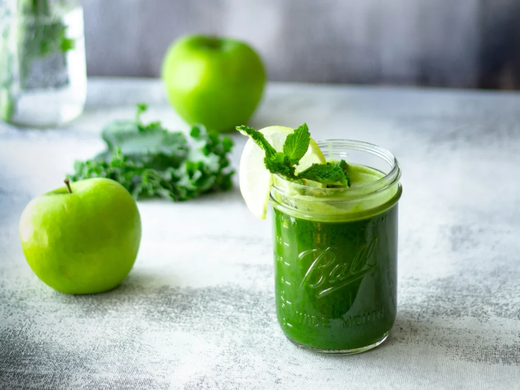 A glass jar of green juice detox garnished with mint leaves and a slice of lime, placed alongside fresh green apples and kale on a light textured background.