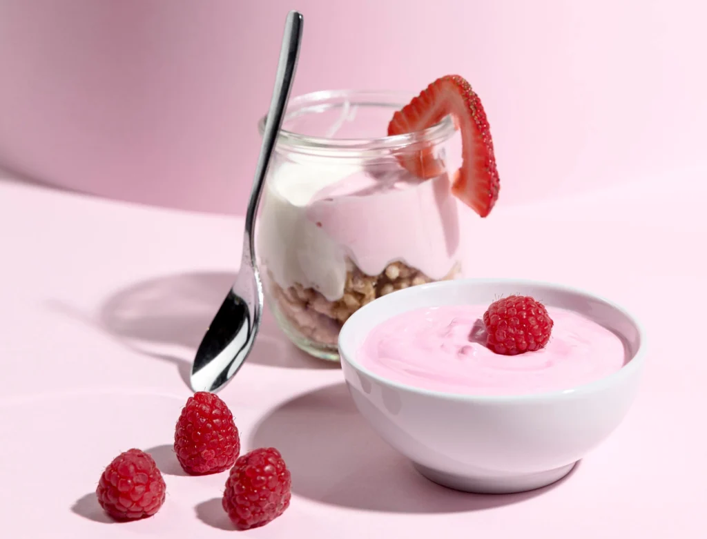 Close-up of pink Greek yogurt smoothie served in a bowl and jar, topped with fresh raspberries and strawberries, on a pastel pink background