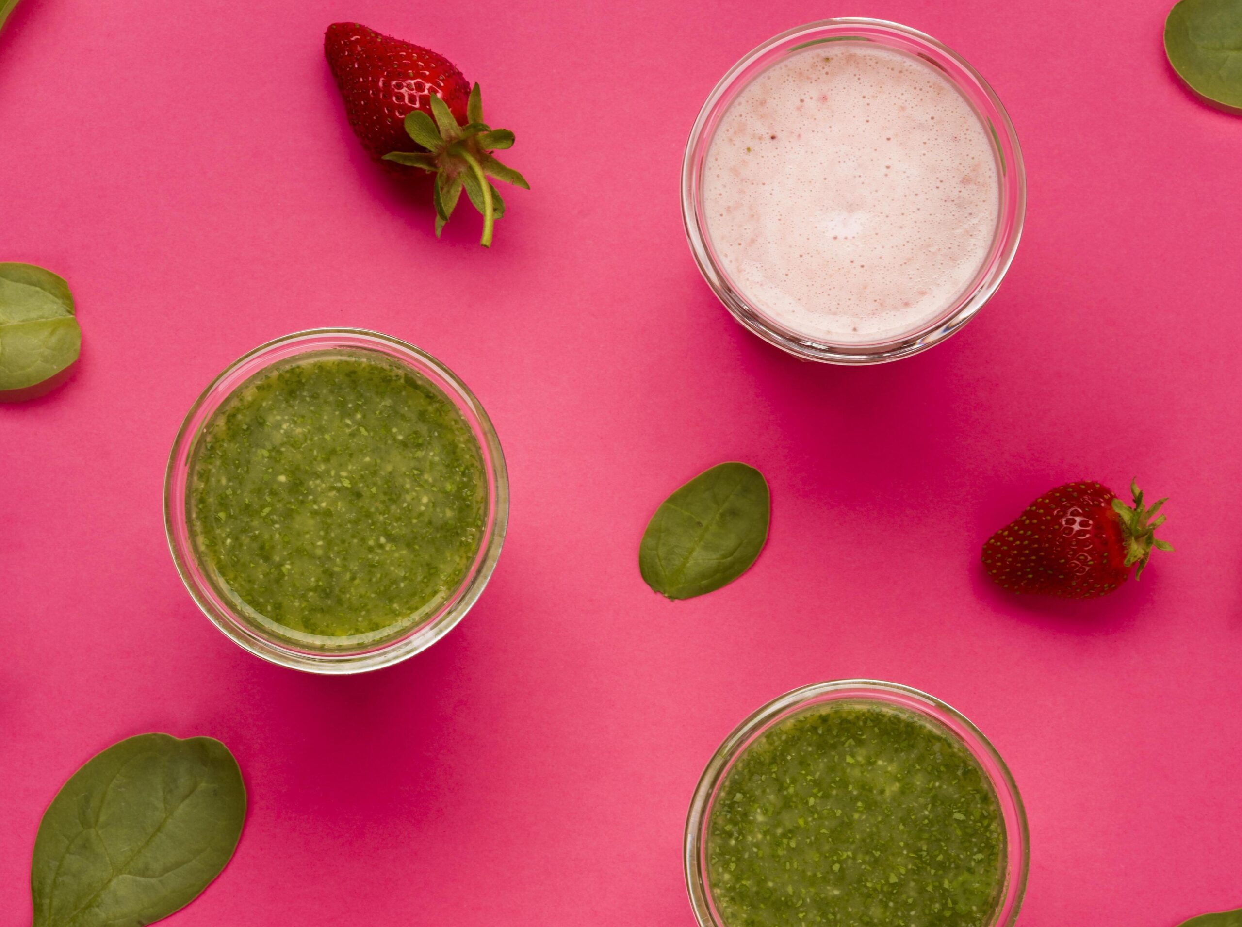 Top view of spinach and berry smoothies in glass cups surrounded by fresh spinach leaves and strawberries on a vibrant pink background.